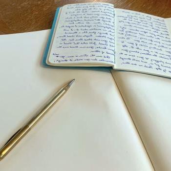 A notebook with writing lays on top of a blank notebook by a pen.
